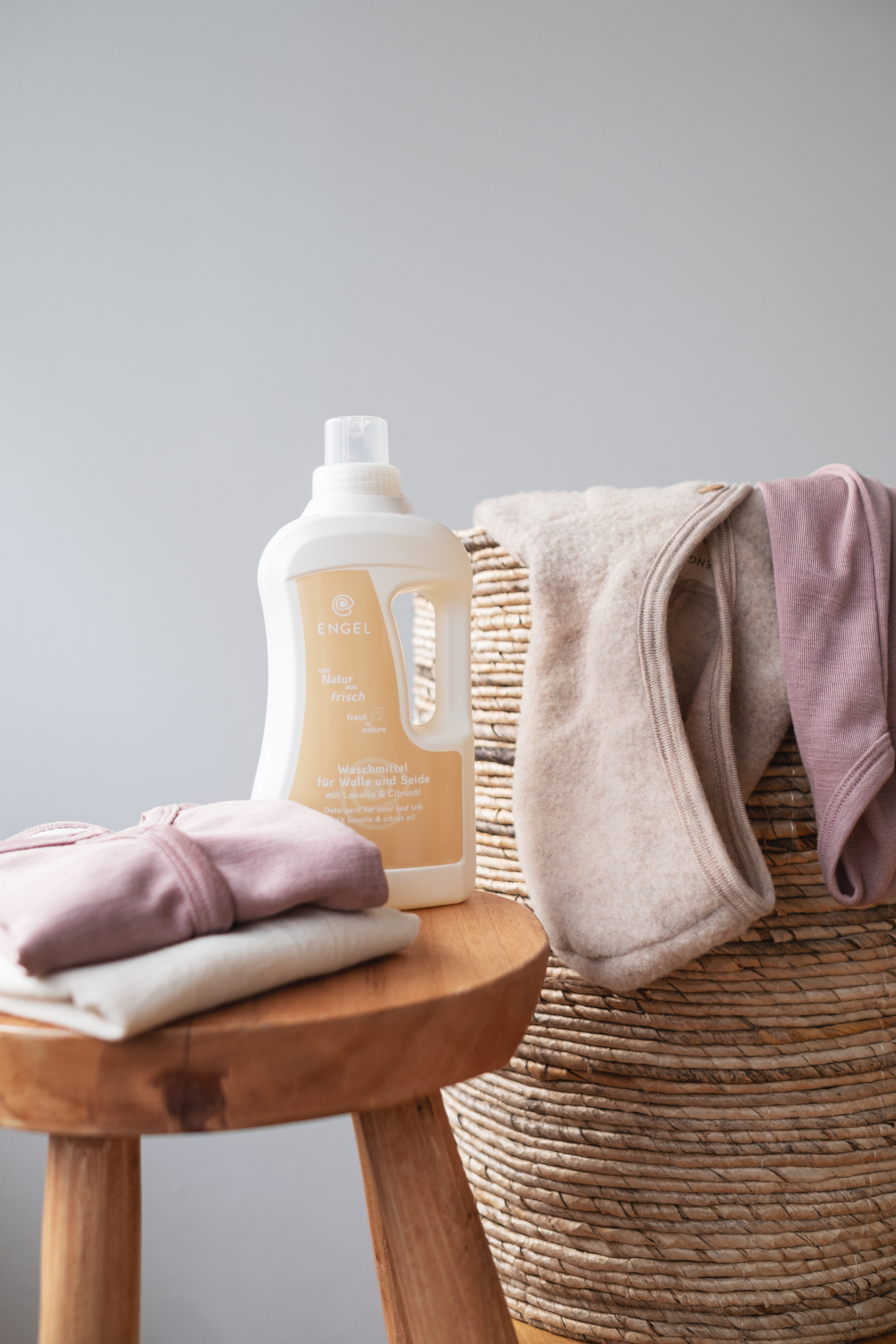 ENGEL Wollwaschmittel für Wolle und Seide neben gefalteter Kleidung und Wäschekorb mit Naturtextilien.